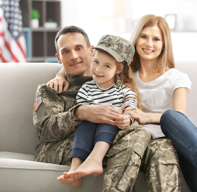 גבר במדי צבא אמריקאי יושב על ספה ומחבק ילדה קטנה בעוד אישה עם חולצה לבנה יושבת לידם ומחייכת ברקע ניתן לראות דגל אמריקאי מטושטש
